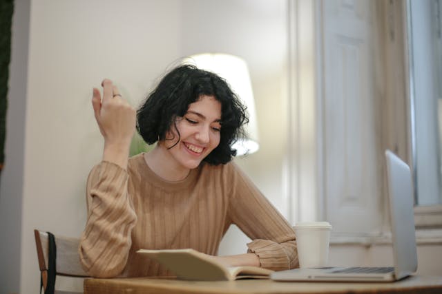 Młoda kobieta z notatnikiem, kubkiem i laptopem. Odniesienie do terapii online i jej skuteczności.
